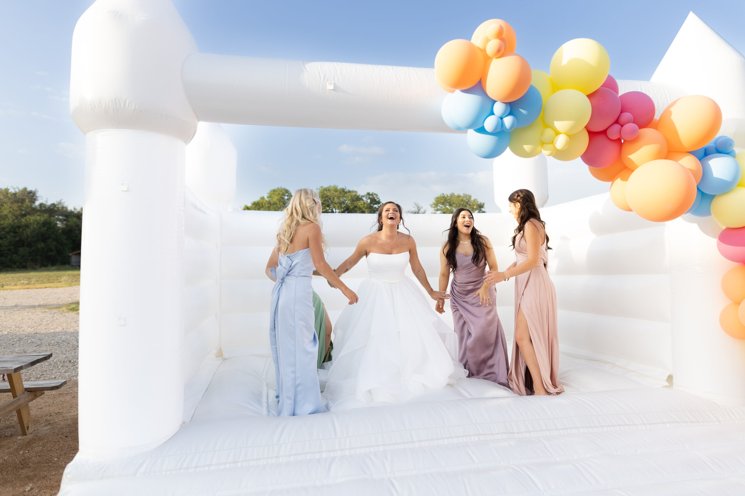 bride and bridesmaids in colorful dresses jumping in a white bouncehouse